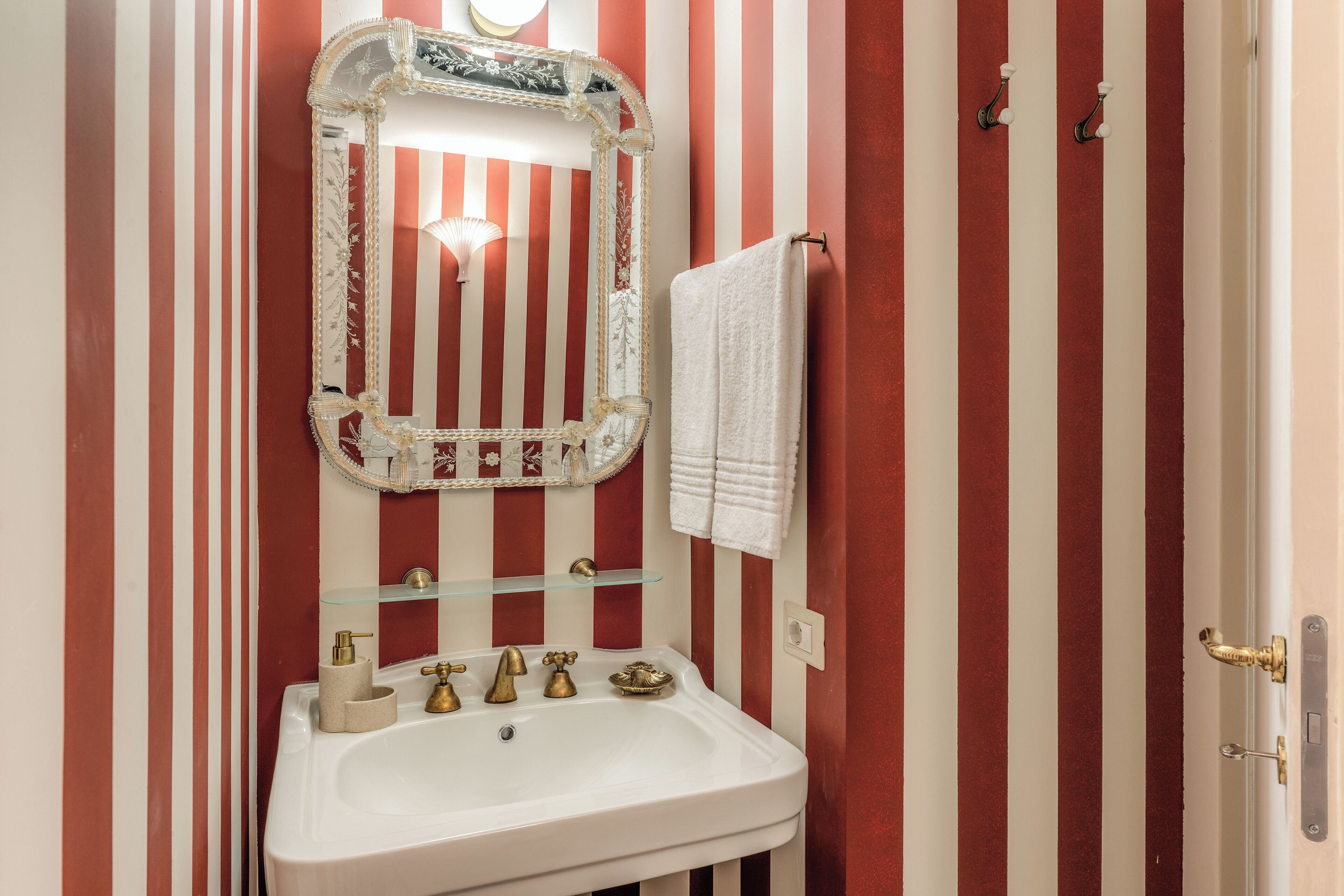 Un baño con un lavamanos blanco, grifos dorados, un estante de vidrio y una pared a rayas rojas y blancas.