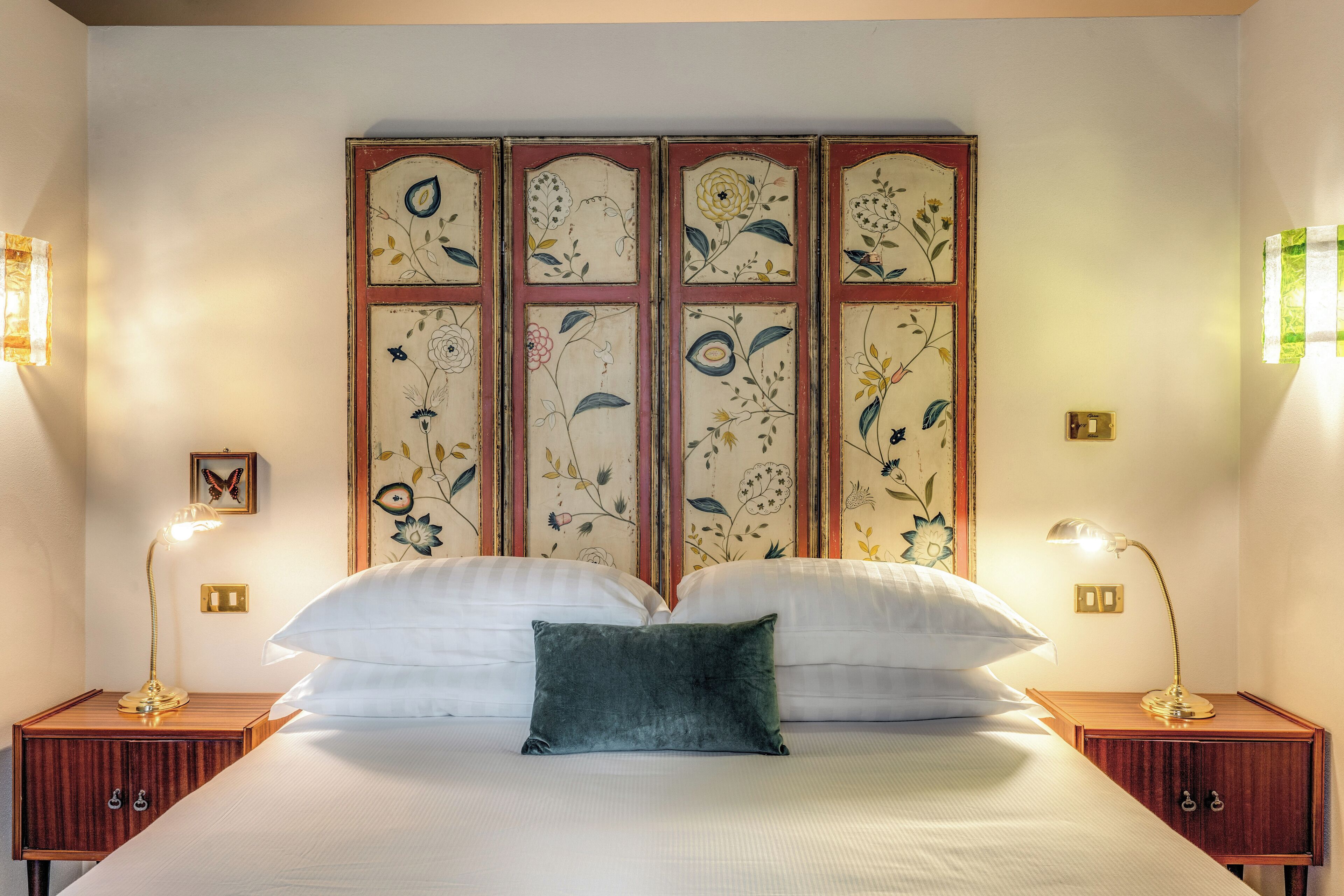 A neatly made bed with white linens and a single green pillow, flanked by two bedside tables with lamps, against a wall with a floral-patterned screen.