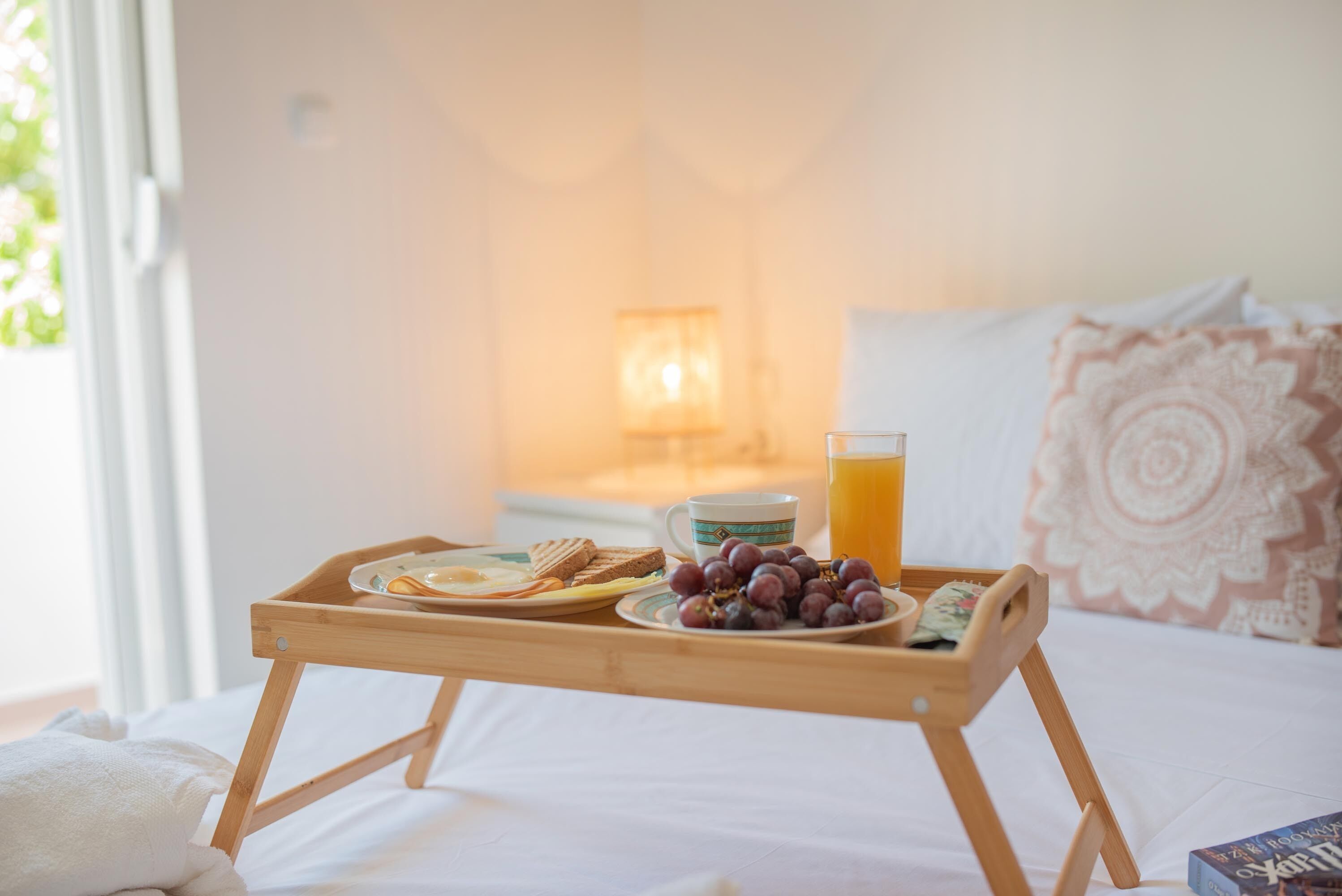 A wooden tray with breakfast items including toast, eggs, and grapes, placed on a bed with a view of a lamp and a patterned pillow.