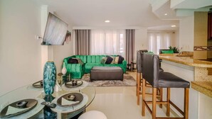 A modern living room with a green sofa, a glass dining table, and a kitchen area with a bar counter.