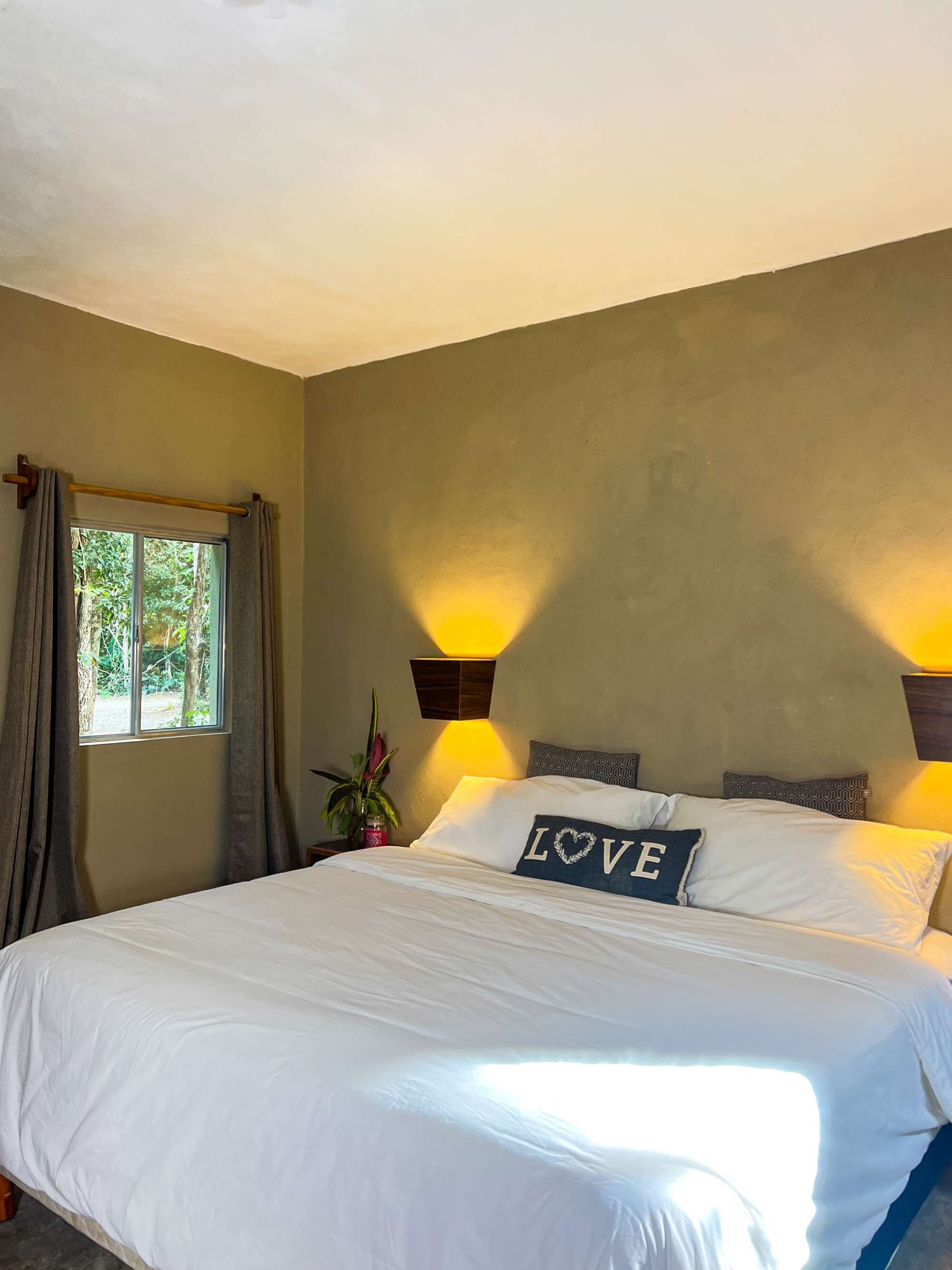 Un dormitorio con cama, ventana con cortinas, lámpara de pared y almohadón decorativo con la palabra 'LOVE'.