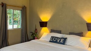 Un dormitorio con cama, ventana con cortinas, lámpara de pared y almohadón decorativo con la palabra 'LOVE'.