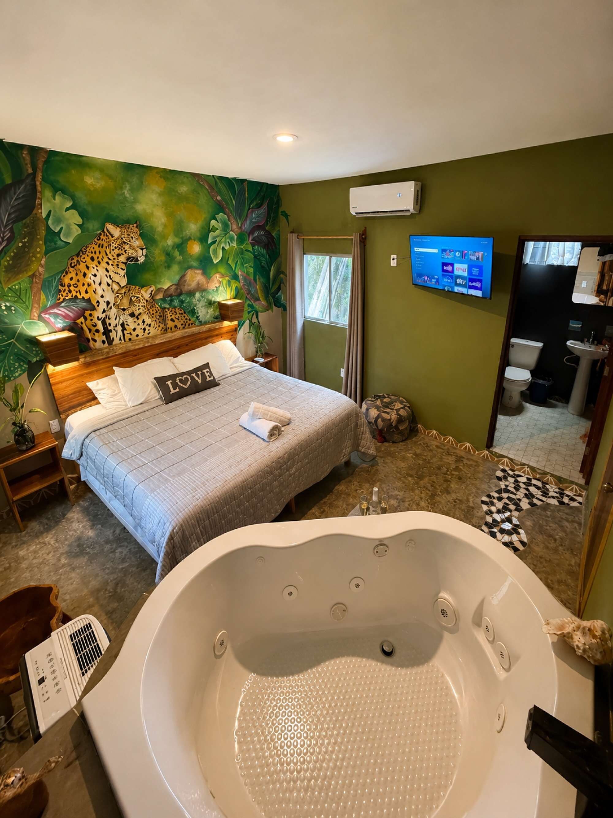 A bedroom with a large bed, a wall mural of a leopard, a television, and a bathroom with a bathtub.