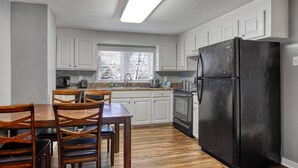 A kitchen with white cabinets, a black refrigerator, wooden chairs, and a window with blinds. - The Cozy Cottage of Florence!

 (Florence)