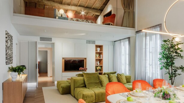 A modern living room with a high ceiling, wooden beams, a dining area with orange chairs, and a TV mounted on the wall. - The Victory Penthouse (Lucca)