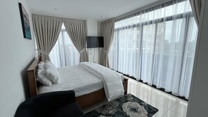 Ein modernes Schlafzimmer mit einem Bett, einem Flachbildfernseher und großen Fenstern mit transparenten Vorhängen.