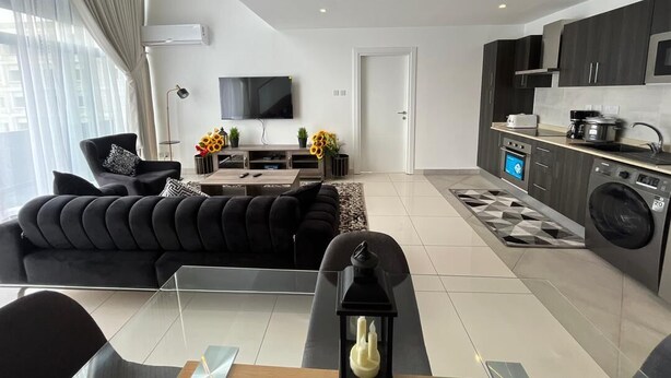 Ein modernes Wohnzimmer mit einer schwarzen Couch, einem Glastisch und einem Flachbildfernseher. Die Küche verfügt über dunkle Schränke, eine Waschmaschine und eine geflieste Spritzschutzwand.