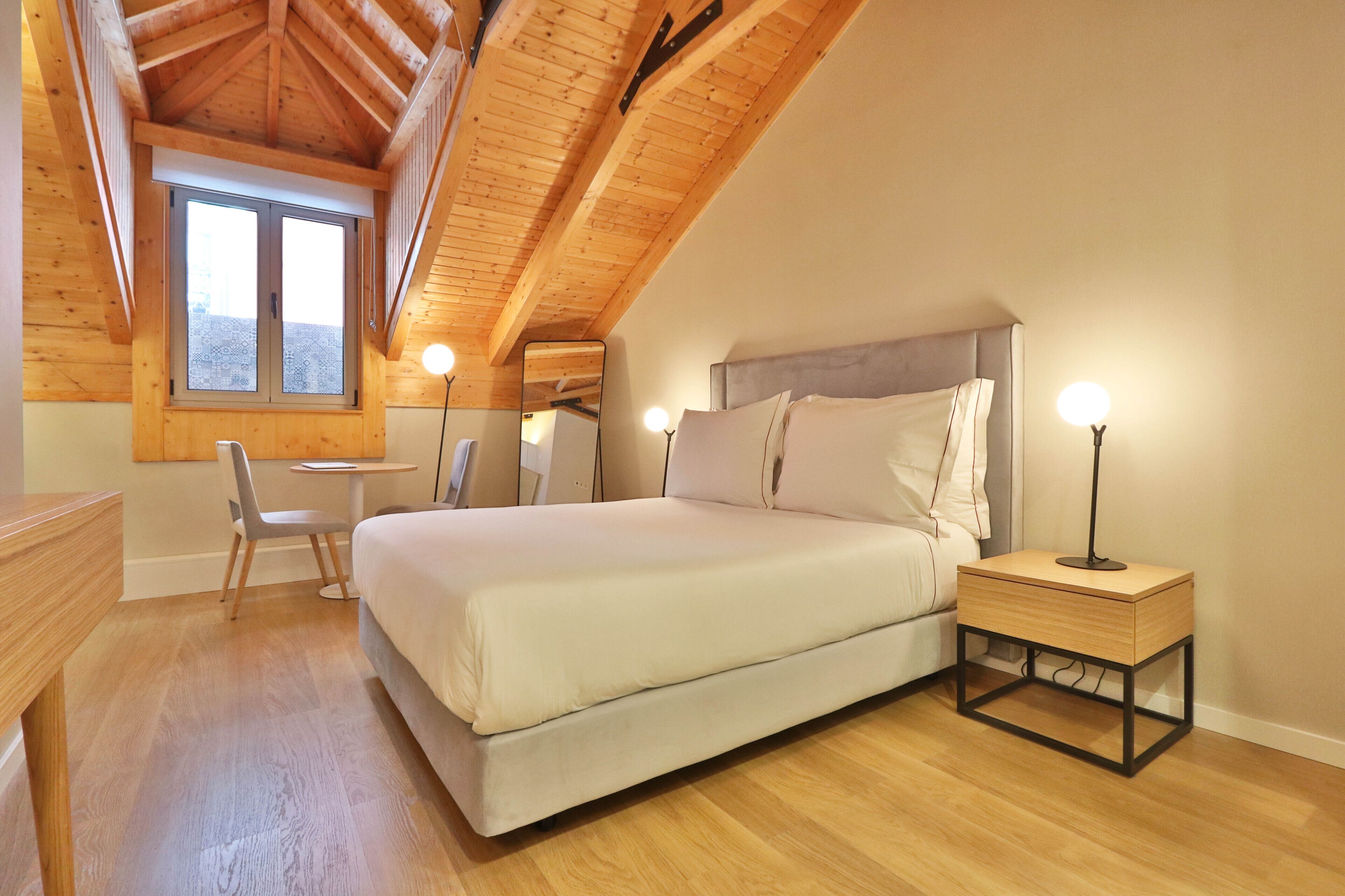 A bedroom with a wooden ceiling, a bed, bedside lamps, a chair, and a small table.