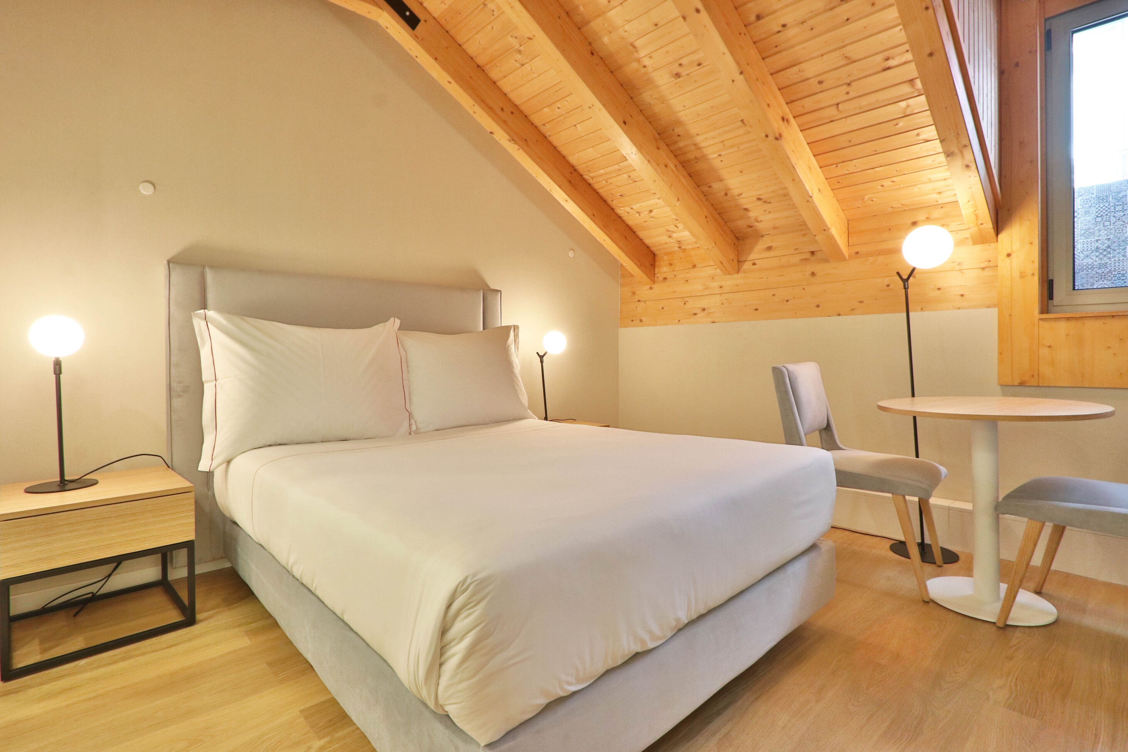 Un dormitorio con cama, mesitas de noche, una silla, una mesa y una ventana.