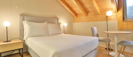 Un dormitorio con cama, mesitas de noche, una silla, una mesa y una ventana.