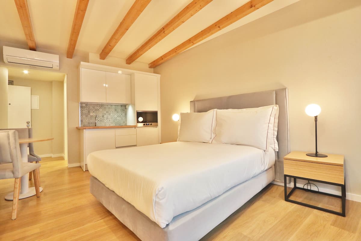 A bedroom with a large bed, bedside table with a lamp, a small desk, and a kitchenette in the background.