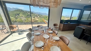 A dining area with a wooden table set for six, large windows offering a mountain view, and a kitchen area with dark cabinetry. - Villa A Trinita: Private paradise, infinity pool, gym and games room (Porto-Vecchio)