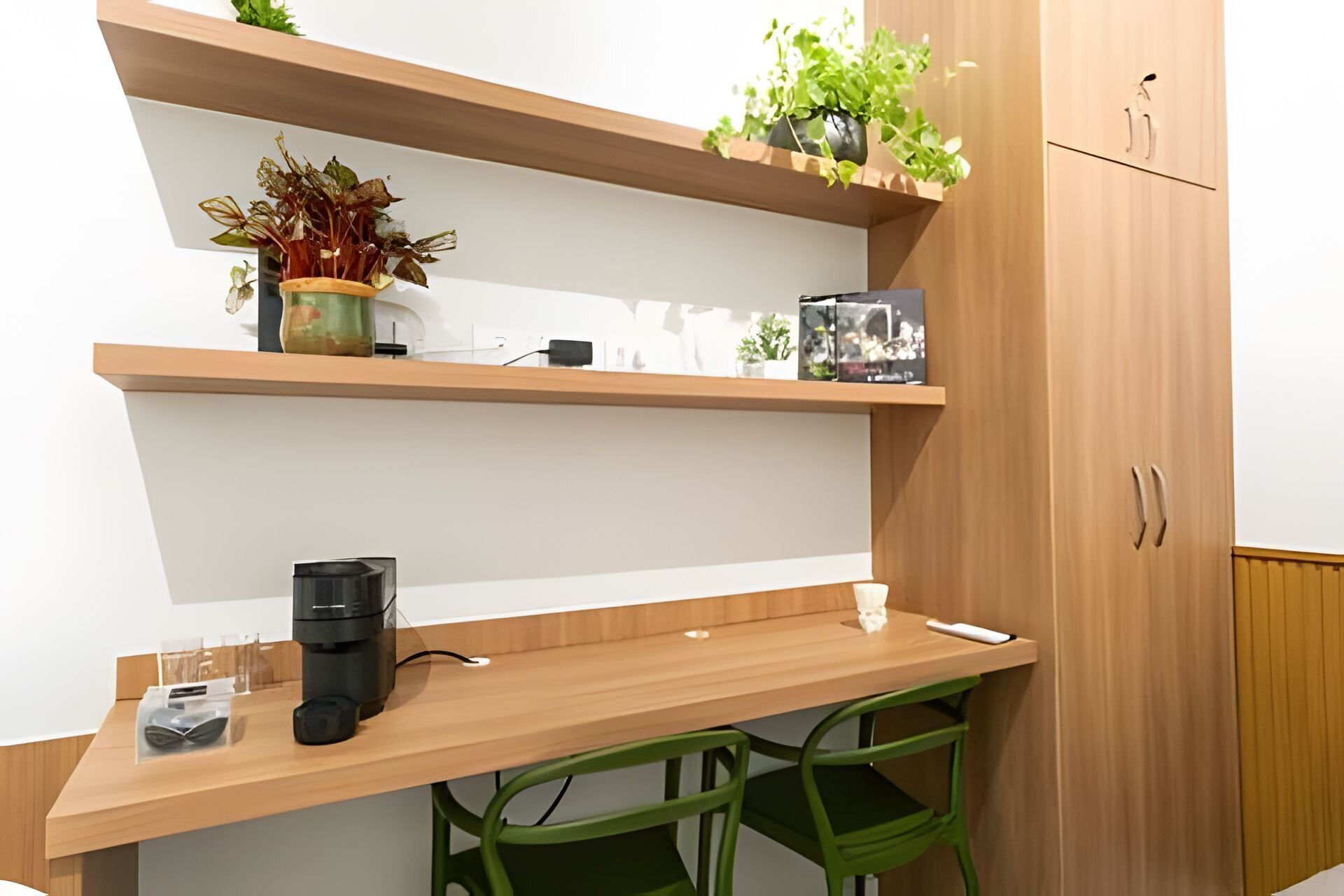 A modern workspace with a wooden desk, a coffee maker, and green chairs. There are two shelves with plants and decorative items.