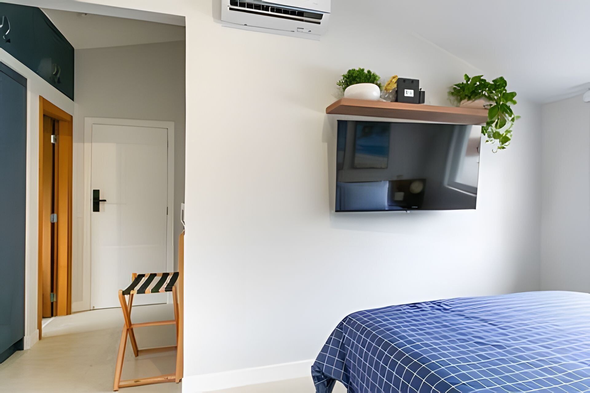 A bedroom with a bed, a TV mounted on the wall, a small shelf with plants, and a door leading to another room.