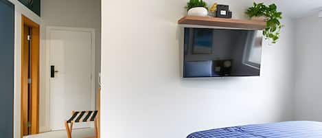 A bedroom with a bed, a TV mounted on the wall, a small shelf with plants, and a door leading to another room.