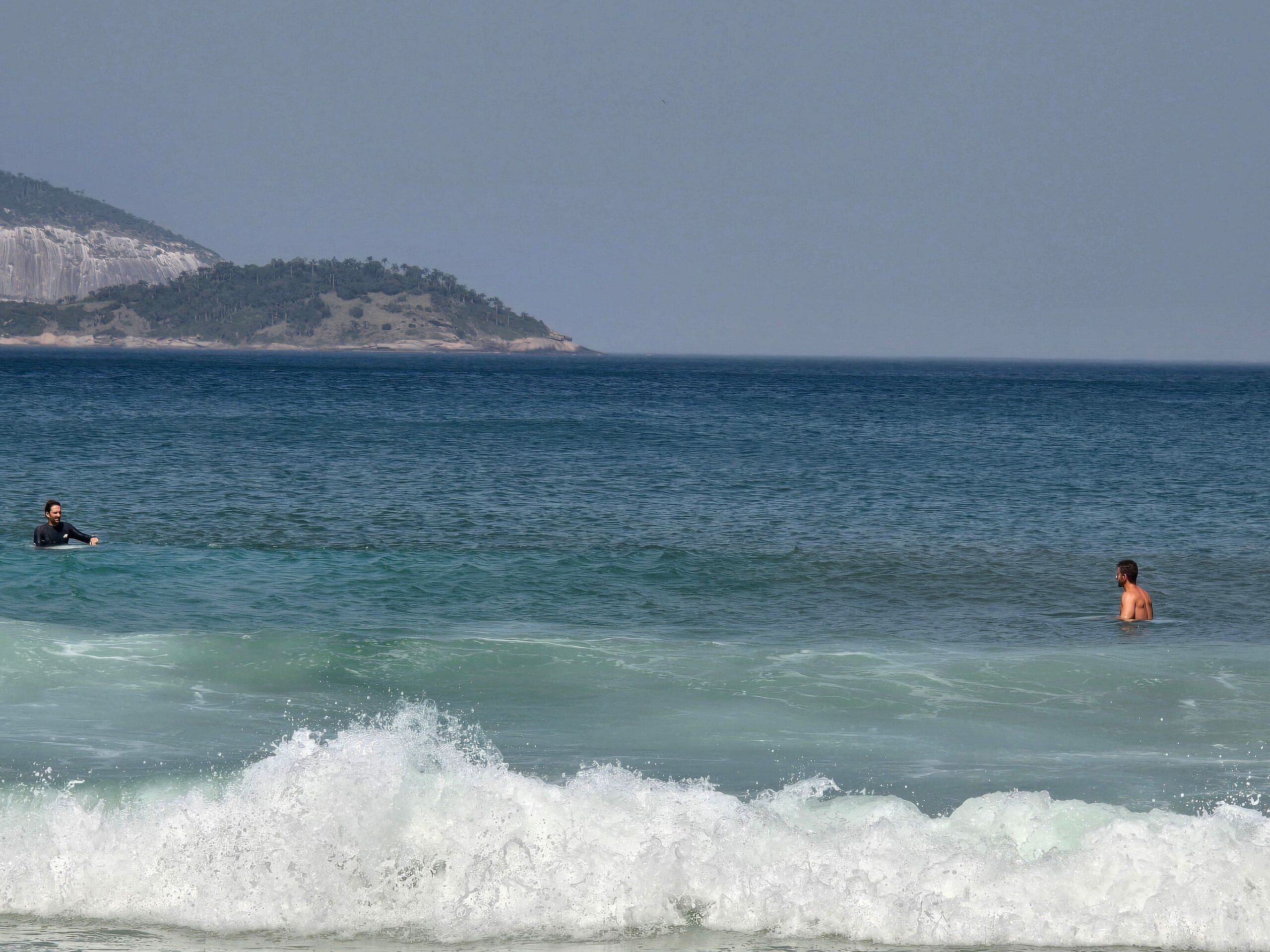 Una playa con dos personas en el agua, cielo despejado y una colina distante.