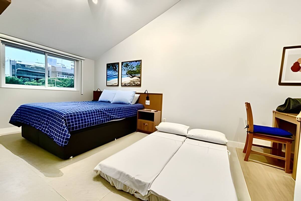 A bedroom with a bed, bedside table, chair, and a desk. There is a window with a view of buildings and greenery.
