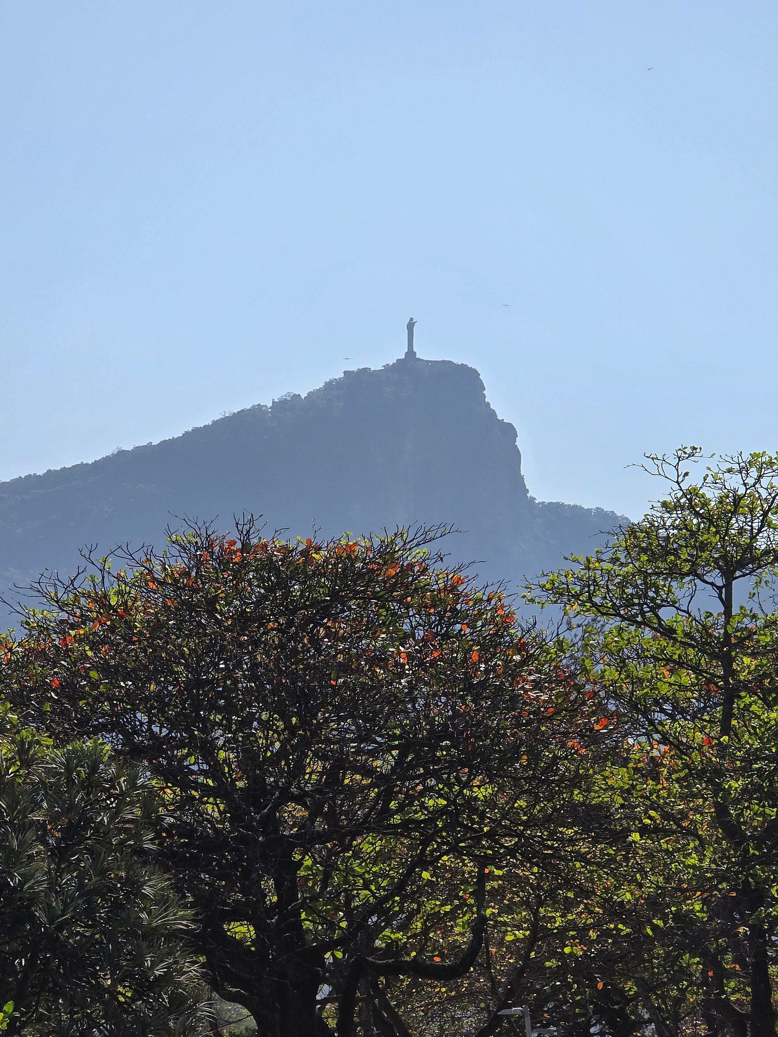 A mountain with a statue on top, surrounded by trees with green and orange leaves.