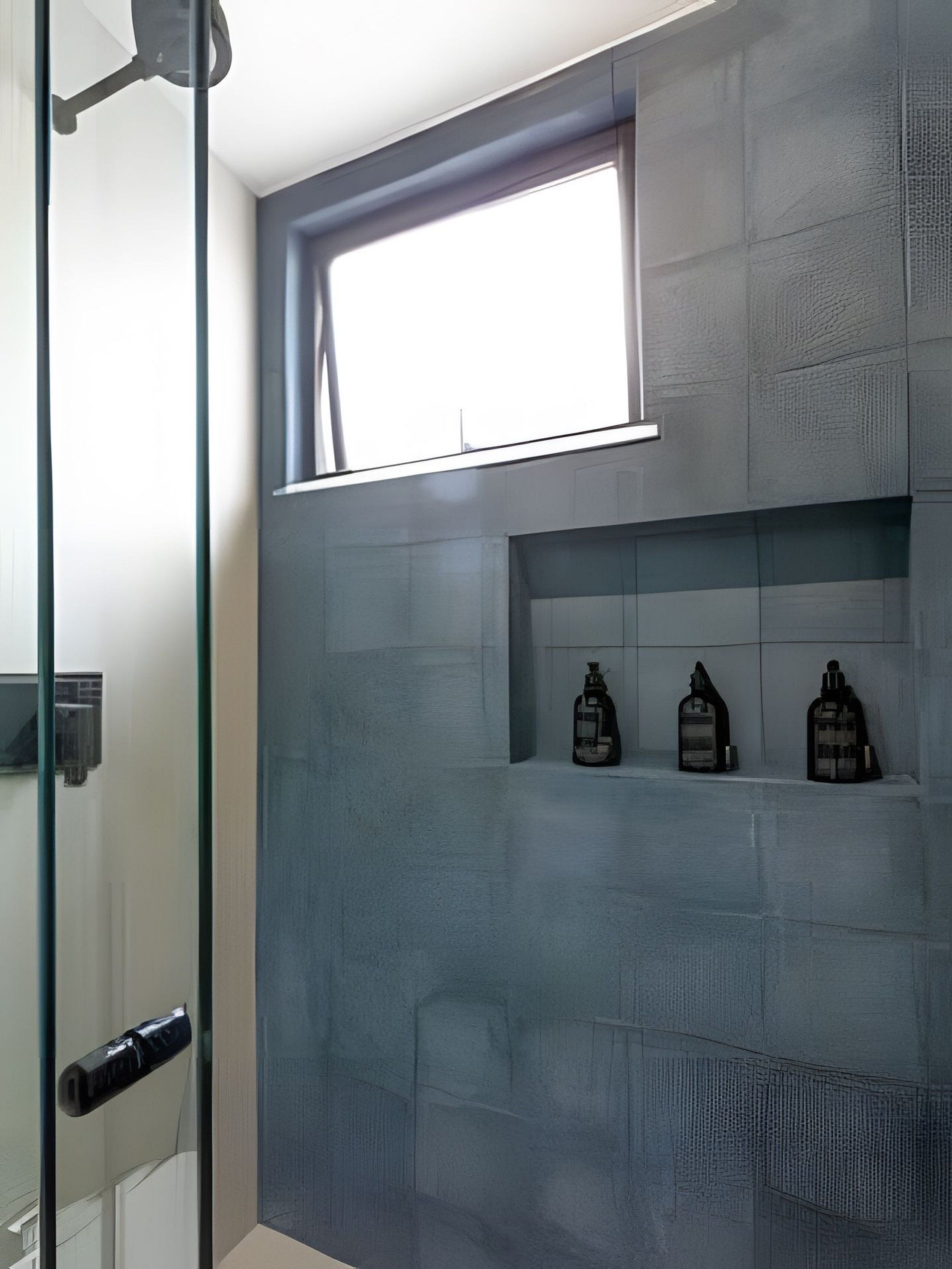 Un baño moderno con cabina de ducha de vidrio, ventana integrada y tres botellas de jabón de manos sobre una repisa.