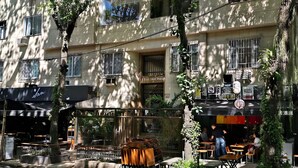 A building with multiple windows, some with metal grilles, and a sign that reads "EDIFICE 5 IRMAOS". There are outdoor seating areas with tables and chairs, and a tree with hanging plants. - chale leblon (Rio de Janeiro)