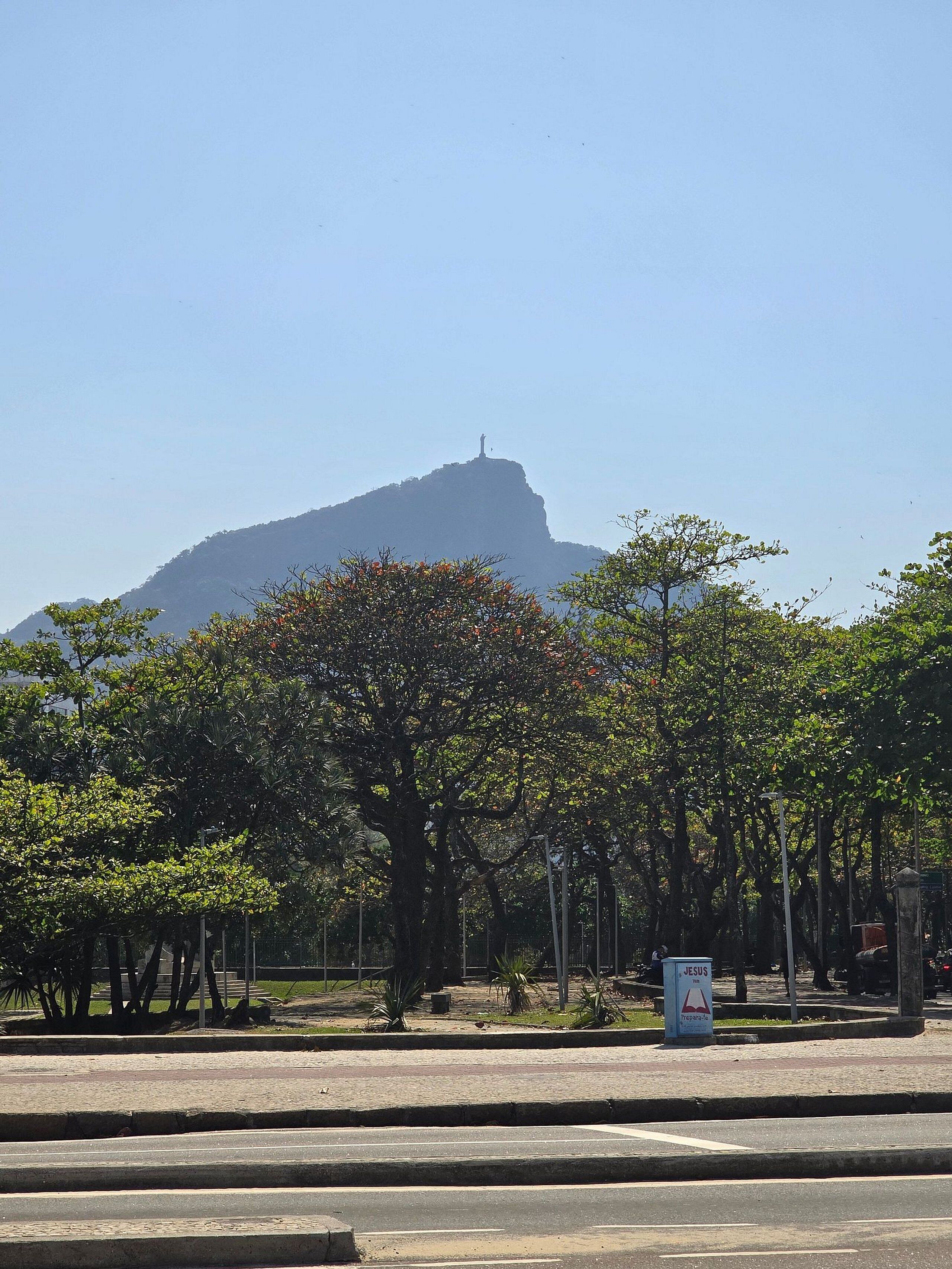 A street view with a prominent hill in the background, trees lining the road, and a sign with the word "JESUS".