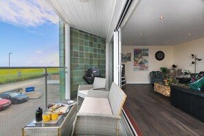 A balcony with a view of a road and fields, a grill, wicker furniture, a sofa, and a poster on the wall.