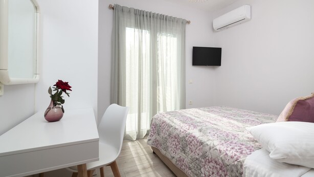 Ein Schlafzimmer mit einem Bett, einem Schreibtisch, einem Sessel, einer Vase mit Blumen, einem Fernseher und einer Klimaanlage.