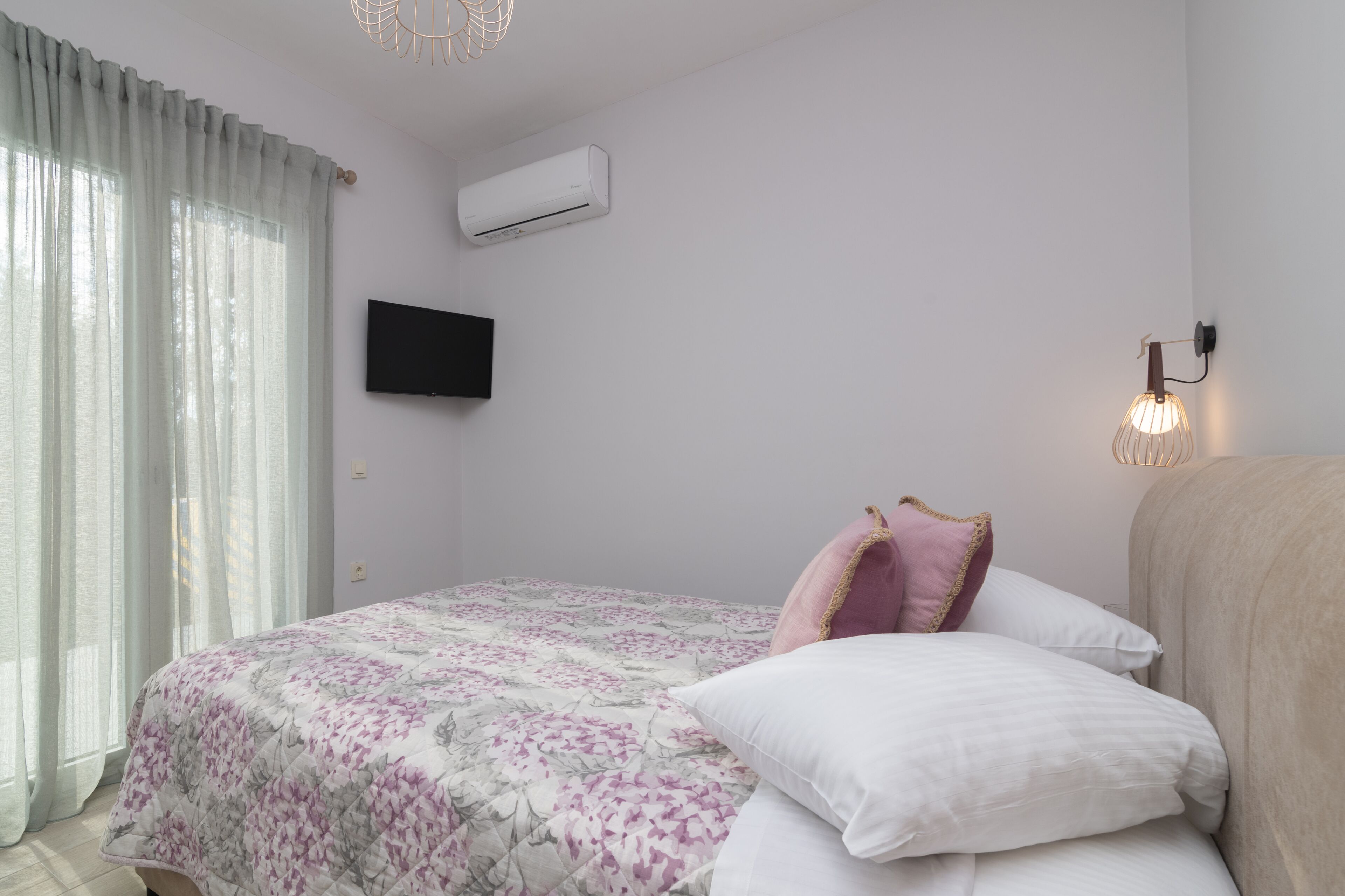 A bedroom with a bed, a flat-screen TV, an air conditioner, and a wall-mounted lamp.