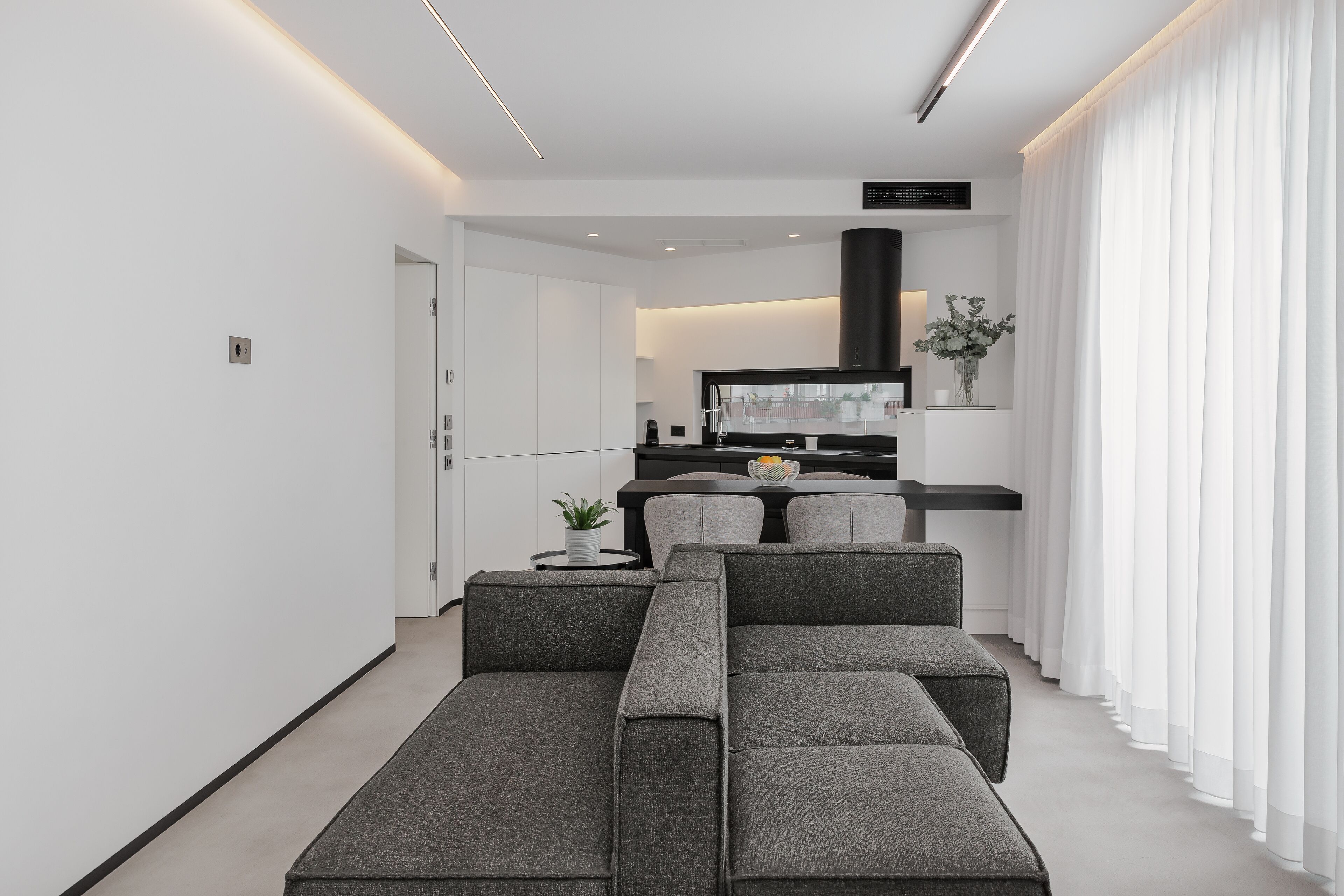 A modern living room with a grey sectional sofa, a flat-screen TV, and a kitchen area in the background.