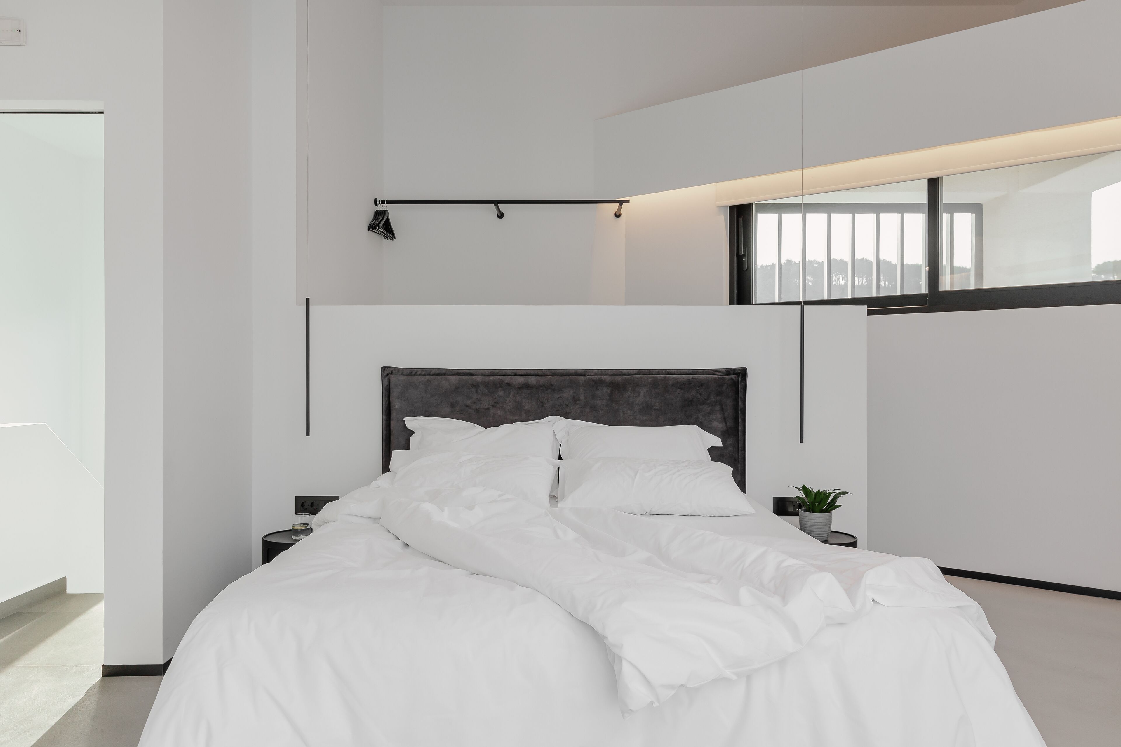 A modern bedroom with a large bed, a bedside table, a small plant, and a window with a view.