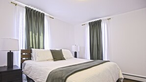 Una cama bien hecha con edredón blanco y manta gris, una lámpara de noche y una mesita de noche con lámpara.