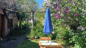 Une terrasse avec une table et des chaises, un parasol bleu et un jardin où les fleurs sont en pleine floraison.