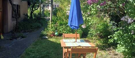 A patio with a table and chairs, a blue umbrella, and a garden with blooming flowers.