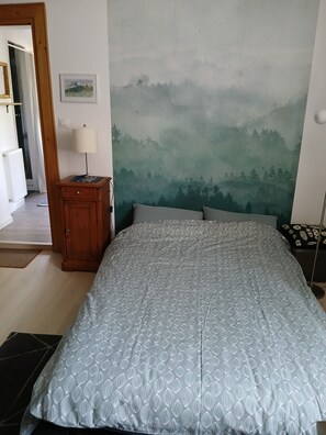 Een slaapkamer met een bed, nachtkastje, lamp, schilderij en uitzicht op een bos.