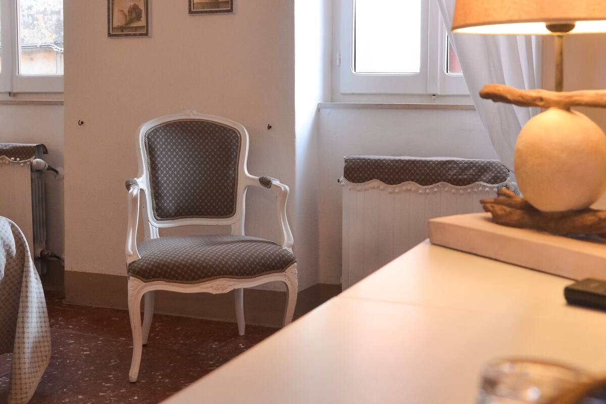 A room with a white chair, a table with a lamp, and a window with curtains. There are framed pictures on the wall.