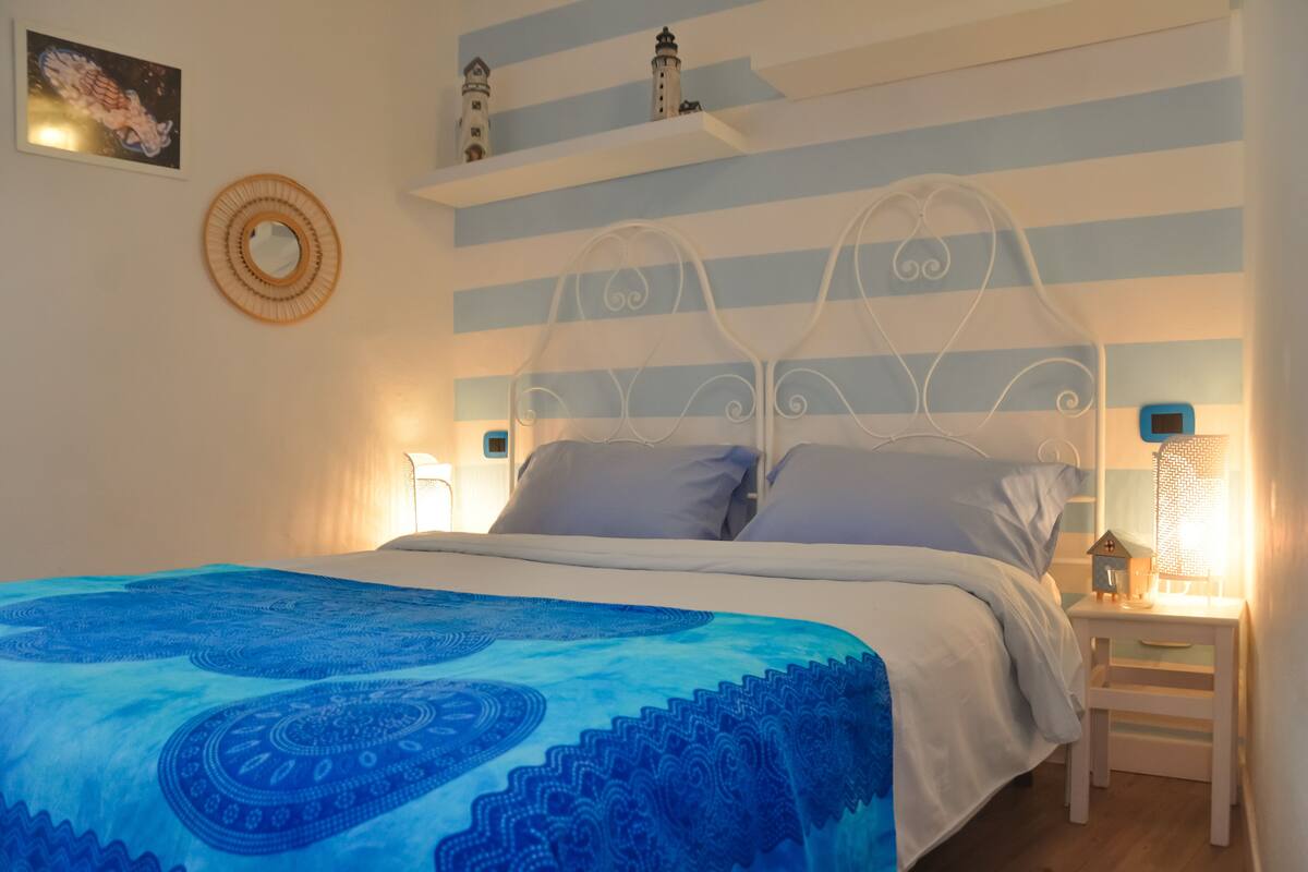 A neatly made bed with a blue patterned bedspread, a white metal headboard, and a small bedside table with a nightlight.