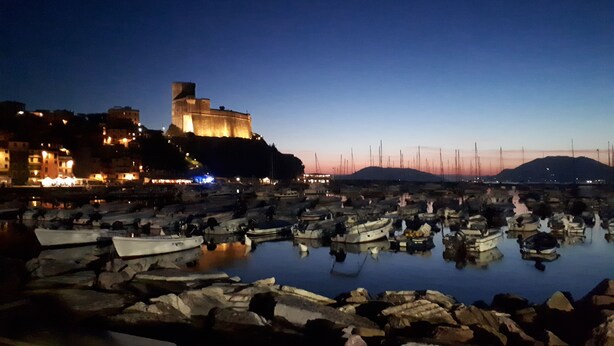 Un port au crépuscule, avec des bateaux amarrés et une structure ressemblant à un château illuminée en arrière-plan.
