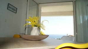 A dining table with a vase of yellow flowers, a wicker tray, and a view of the sea through an open door.
