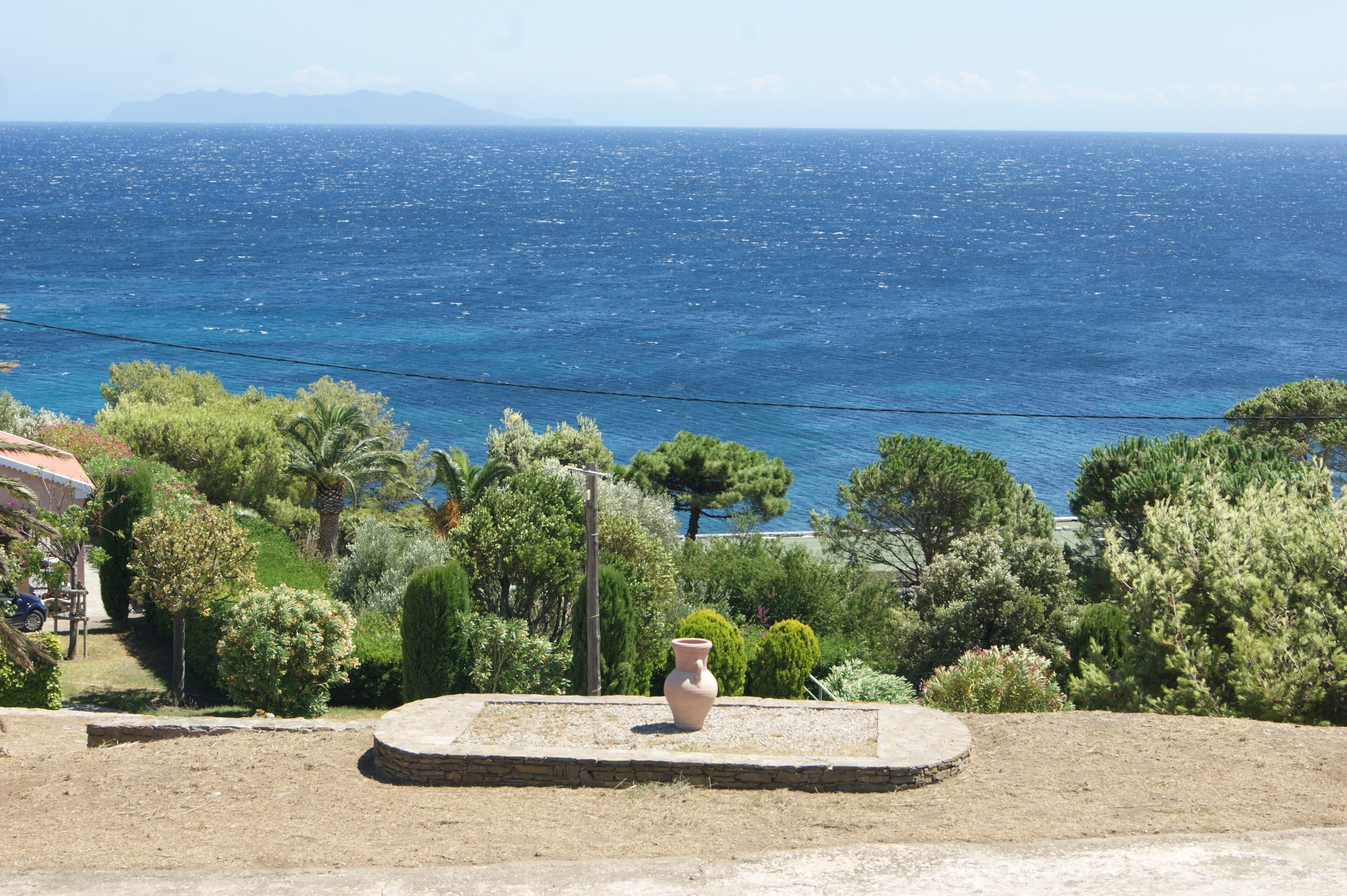A coastal landscape with a view of the sea, a stone bench, and a variety of trees.