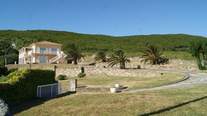 A two-story house with a stone wall and a curved driveway, surrounded by greenery and a hill in the background.