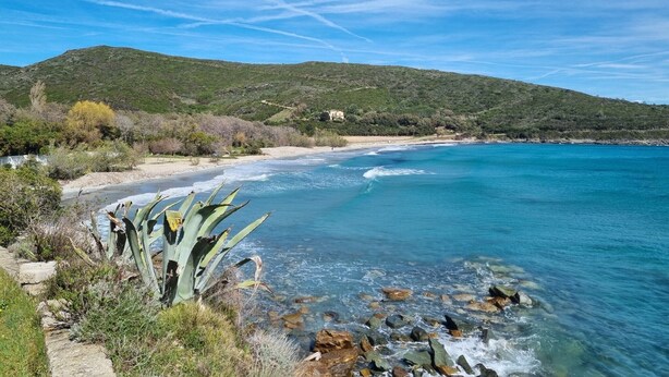 A coastal landscape with clear blue waters, rocky shore, and a hill with sparse vegetation.