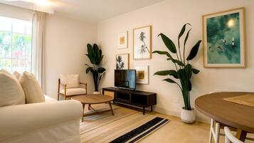 Uma sala de estar moderna com sofá, mesa de centro, televisão e mesa de jantar. Há plantas grandes e quadros nas paredes.