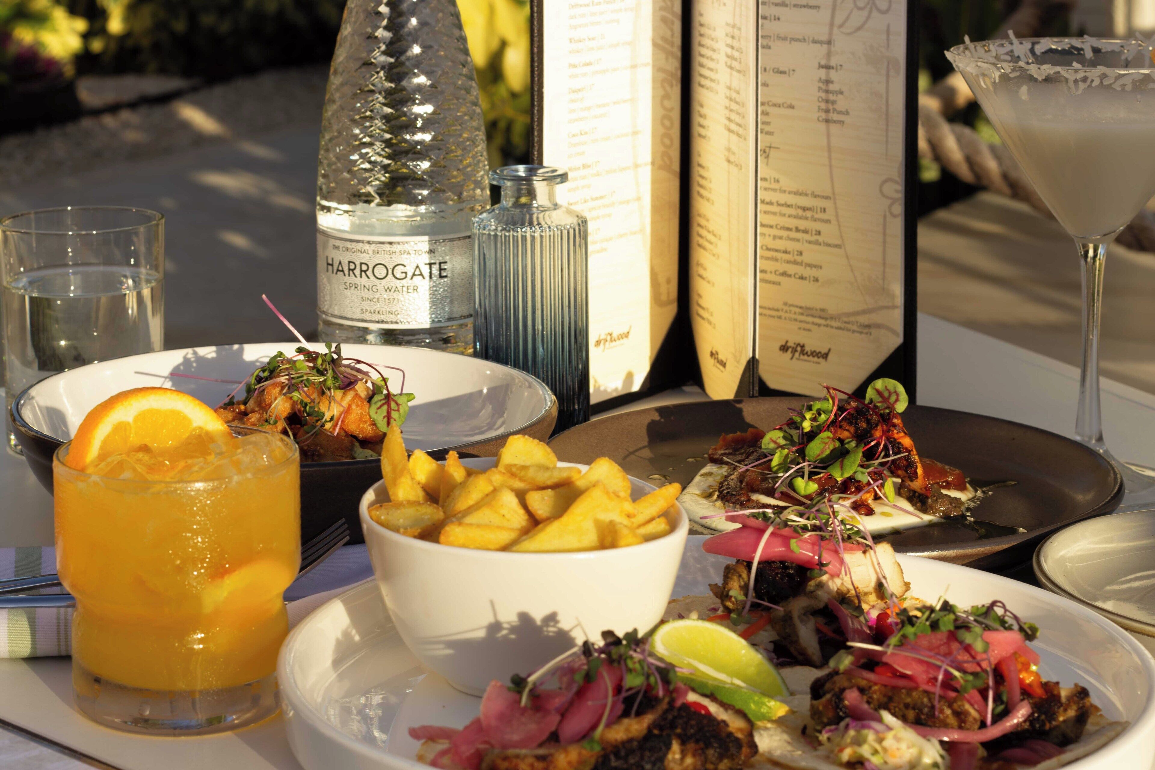 Una mesa con diversos platos, como papas fritas, una ensalada y un plato de pescado, además de una botella de agua con gas Harrogate y una copa de cóctel.