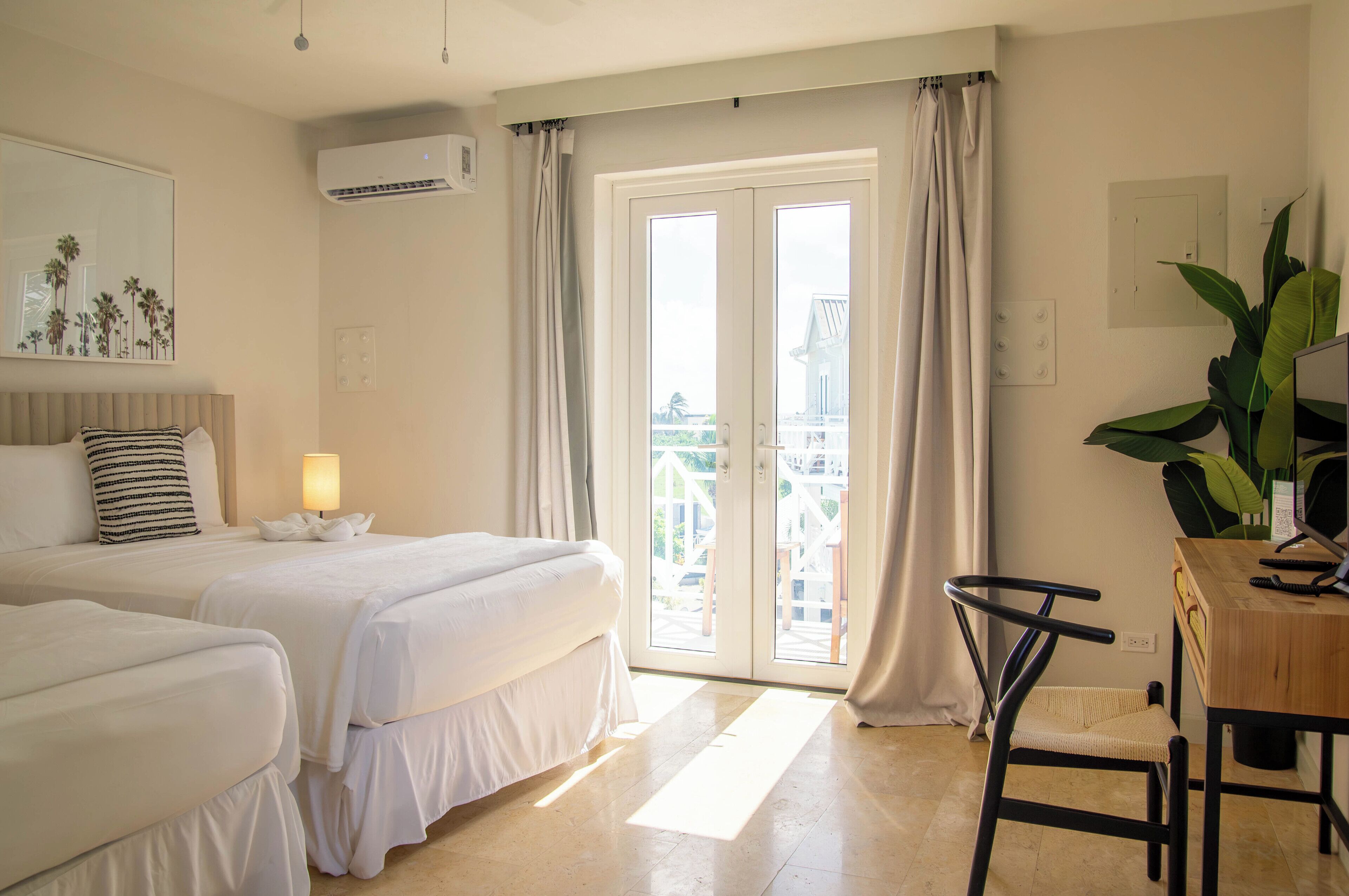 A hotel room with two beds, a desk with a computer, a chair, a lamp, and a large window with curtains.