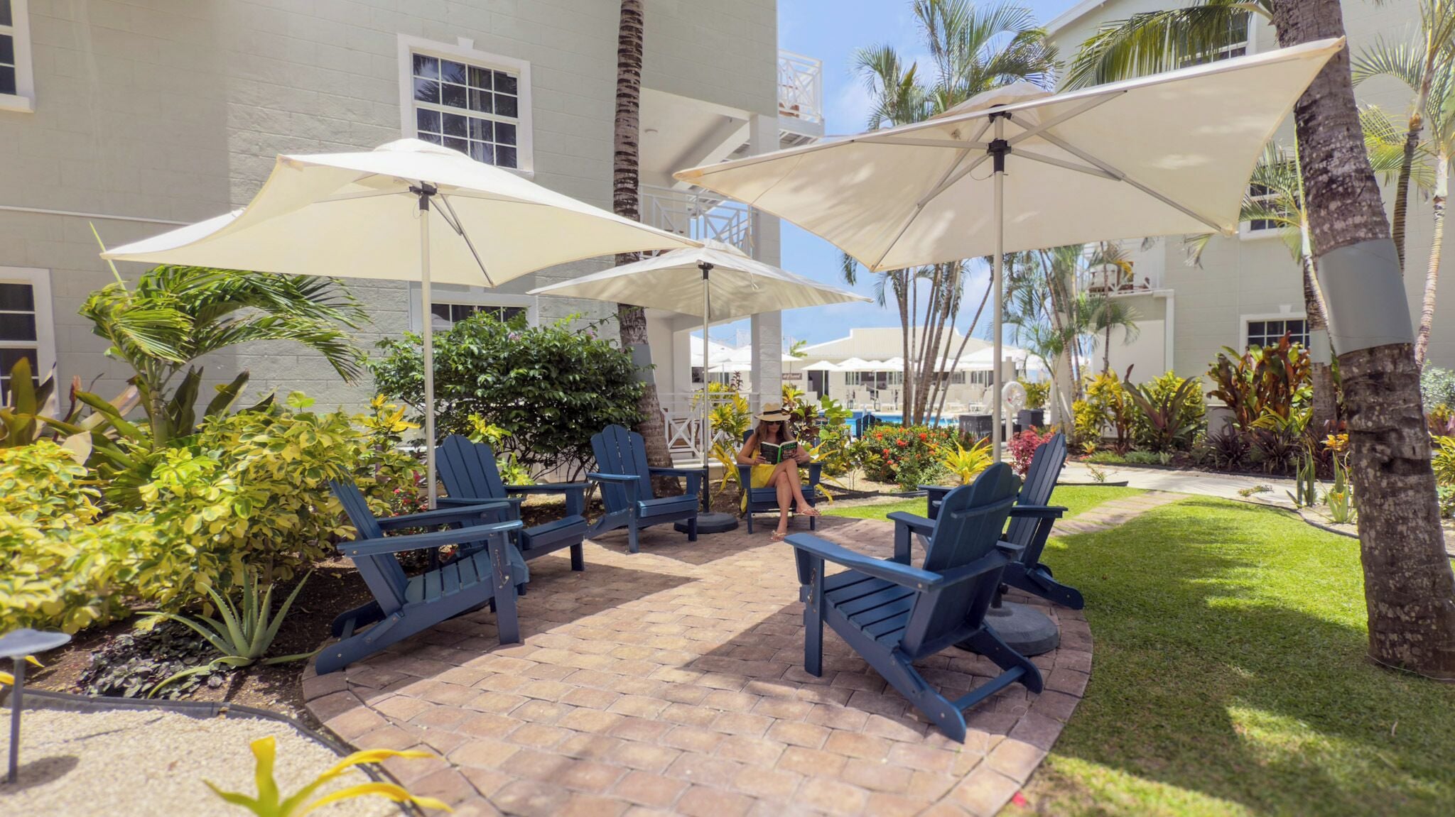 A patio area with blue Adirondack chairs and white umbrellas, surrounded by tropical plants and a pool in the background.