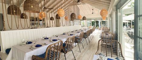 A restaurant with long tables, wicker chairs, and hanging light fixtures.