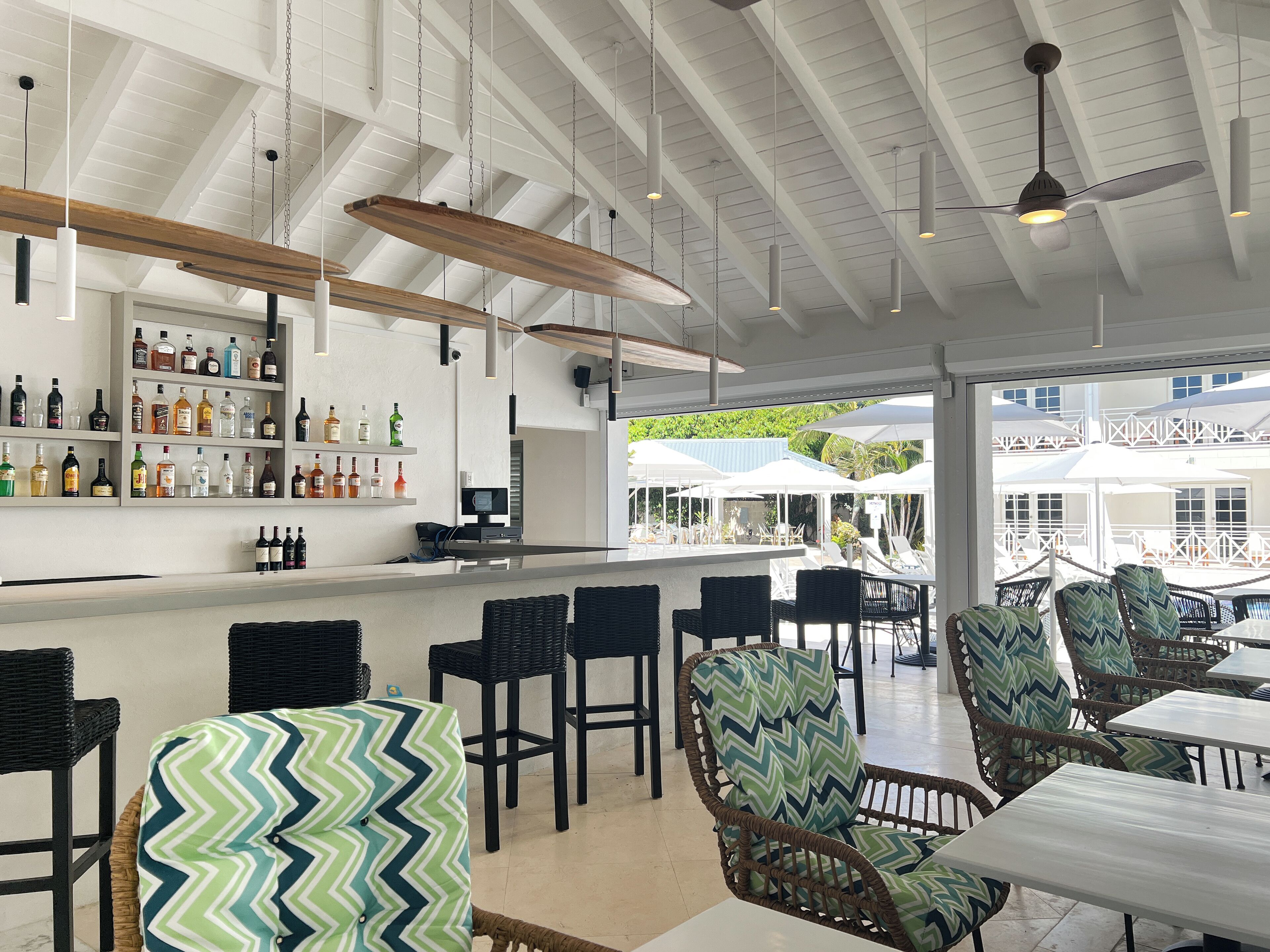 A bar area with a long counter, shelves with bottles, wicker chairs, and a view of outdoor umbrellas and chairs.