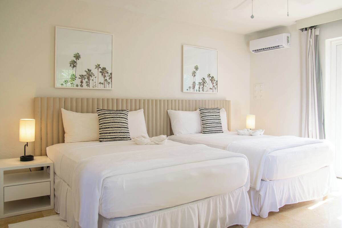 A hotel room with two beds, each with white bedding and striped pillows. There are wall-mounted air conditioning units and two framed pictures of palm trees.