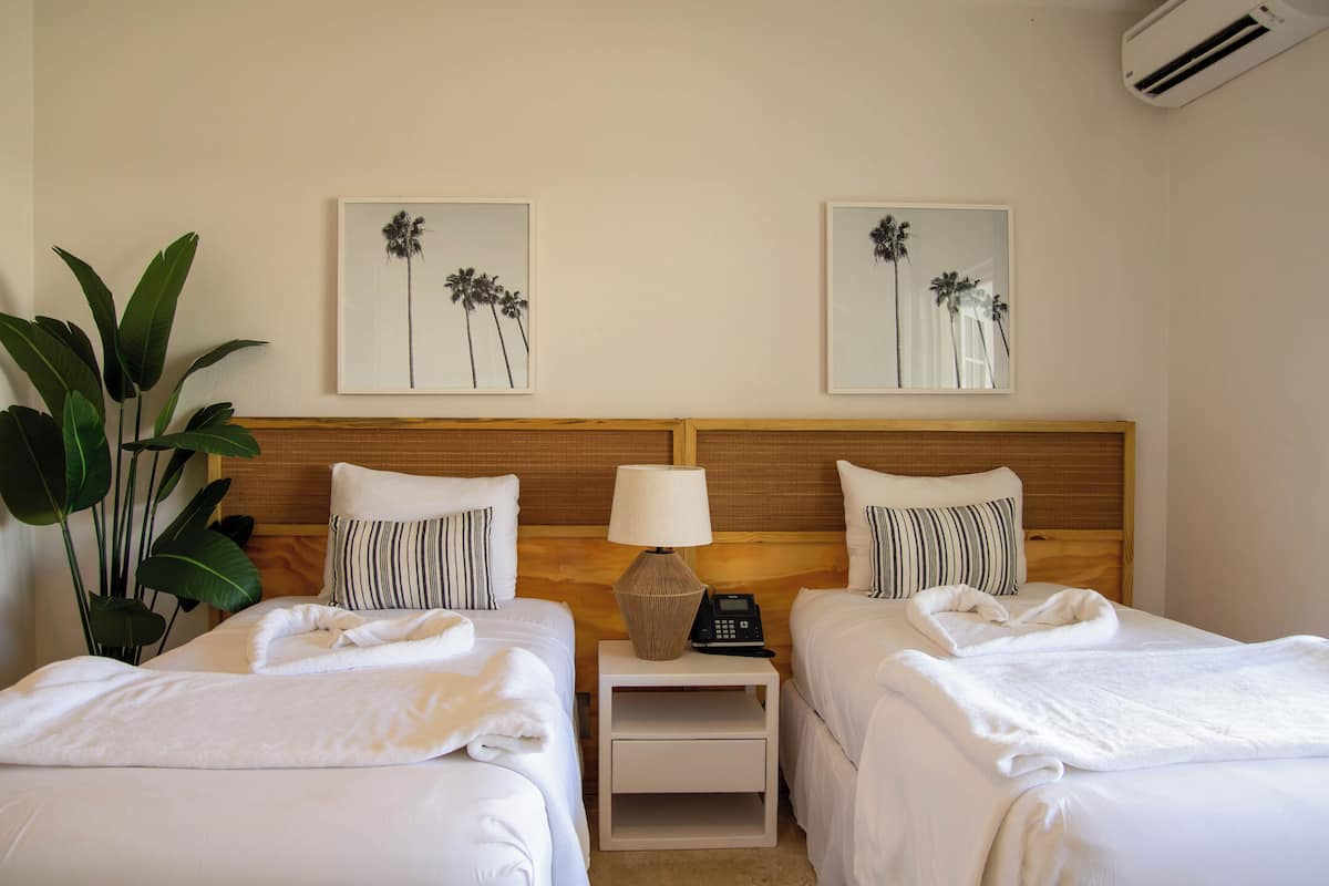 A hotel room with two beds, a bedside table with a lamp, and two framed pictures on the wall.