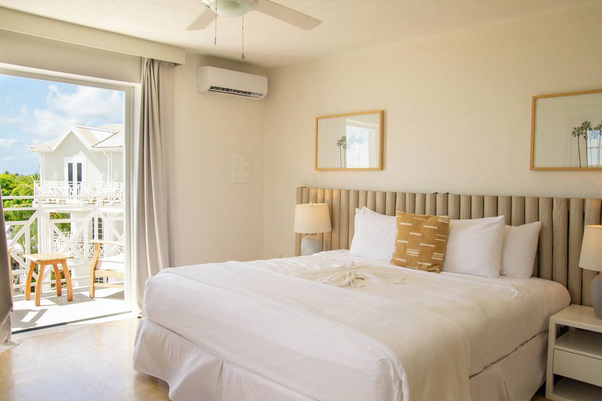 A hotel room with a large bed, two bedside lamps, a ceiling fan, a balcony with a wooden chair, and a view of a white building.